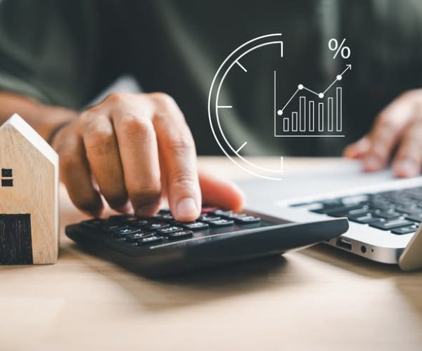 Hand using calculators, planning home refinance. Wooden house model, buy or rent note, calculators on desk. Saving for property purchase, mortgage payment. Tax, credit analysis for financial benefit.