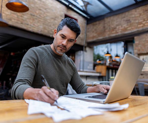 Latin American business manager doing the books at a restaurant using his laptop while writing on a ledger